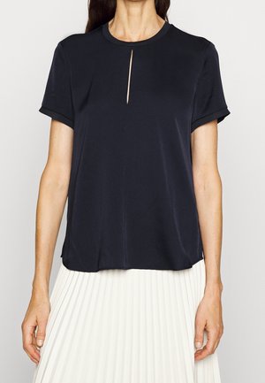 Vrouw die een zwart blouse met korte mouwen en een kleine inkeping bij de hals draagt, gecombineerd met een witte geplooide rok, staand tegen een effen achtergrond.