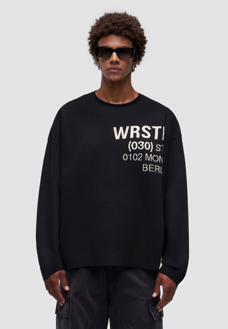 Svart sweatshirt med oversized passform, rund halsringning och långa ärmar. Har vita textgrafiker som inkluderar "WRST" och "BERL" på framsidan.