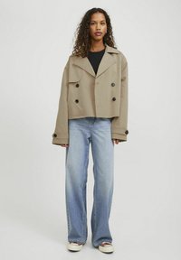 Giacca doppio petto beige tagliata, con grandi bottoni neri, ampi risvolti e maniche larghe. Abbinata a jeans a gamba larga blu chiaro.