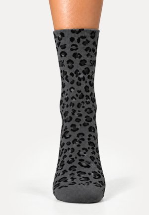 Un pie lleva un calcetín gris de caña media con estampado de leopardo negro sobre un fondo blanco.