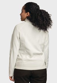 Giacca in pile di colore crema chiaro con collo alto, maniche raglan, dettagli di cucitura sottili e una texture liscia, abbinata a pantaloni scuri.