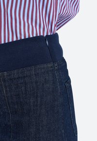 Temno modri jeans z rebrastim pasom in zadrgo. Tkanina ima subtilno teksturo z izrazitim šivanjem in žepi.