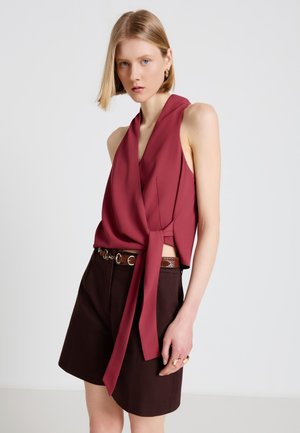 Donna che indossa un top bordeaux senza maniche avvolgente legato in vita e pantaloncini marroni scuro a vita alta con cintura a catena, in piedi davanti a uno sfondo semplice.