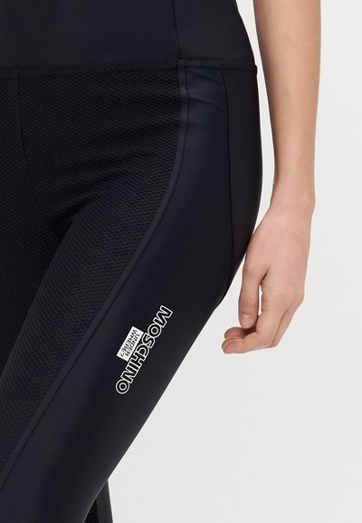 Črne legice z teksturirano mrežasto ploščo in belim logotipom Moschino. Povečan pogled na tkanino in roko na predelu stegna.