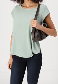 Femme portant un haut à manches courtes vert clair et un jean foncé, tenant un sac à bandoulière en cuir marron foncé sur son épaule droite.