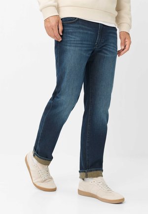 Mann trägt dunkelblaue hochgekrempelte Jeans, cremefarbenen Pullover, weiße Socken und beige Turnschuhe, steht mit einem Fuß leicht nach vorne.