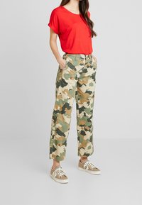 Czerwony top z krótkim rękawem w połączeniu z luźnymi spodniami moro w zielone, beżowe i br�ązowe wzory oraz beżowe trampki z białymi akcentami.