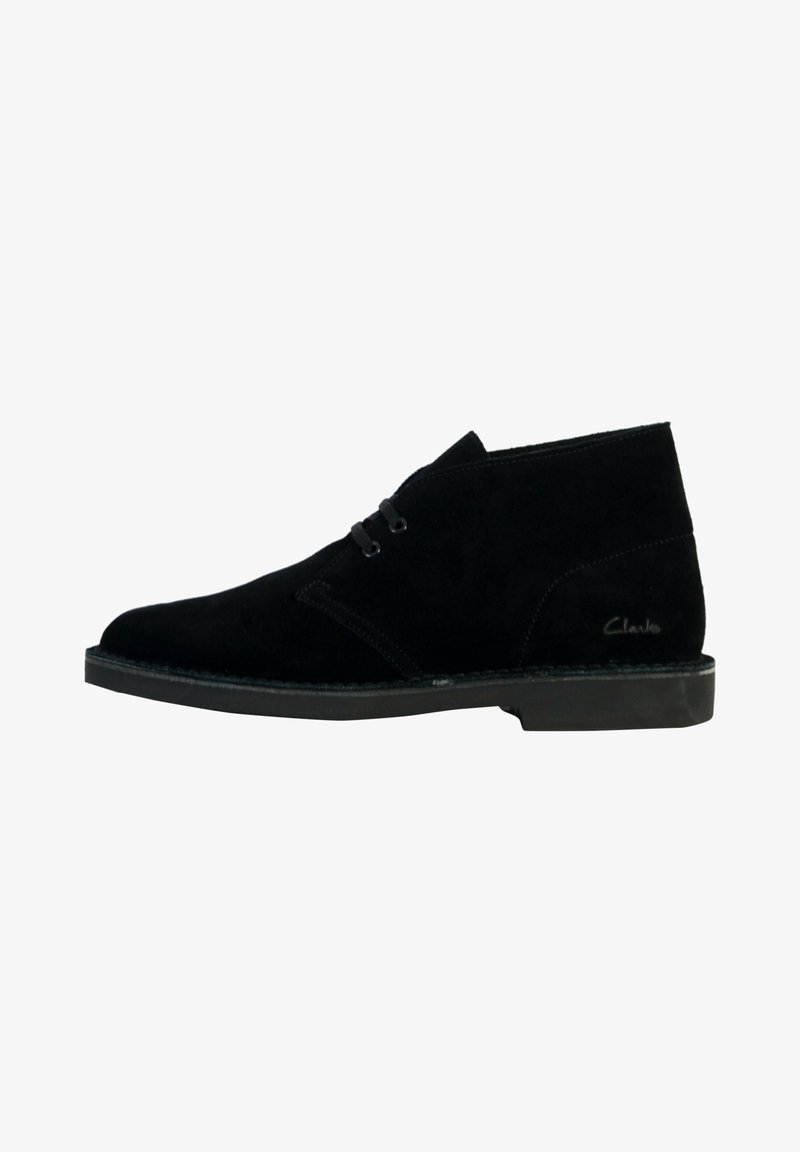 Bottines en daim noir avec deux œillets, semelle plate et coutures visibles sur le côté, conçues pour un usage décontracté ou formel.