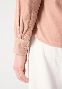 Bras gauche portant une chemise en velours côtelé rose à manches longues avec poignets boutonnés reposant à côté d'un pantalon blanc texturé.