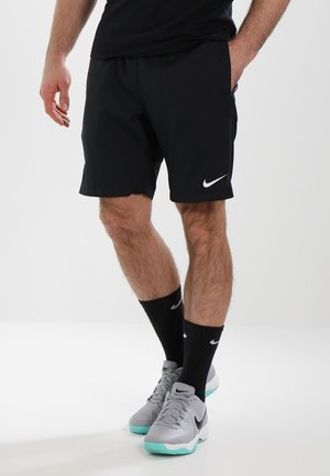 Schwarze Sportshorts mit weißem Nike-Logo, kombiniert mit schwarzen Socken und grauen Nike-Schuhen mit einer hellblauen Sohle. Schlichter Hintergrund.