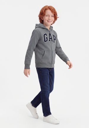 Dreng med rødt hår iført en grå GAP-hoodie, mørkeblå jeans og hvide sneakers, som går og smiler mod en ensfarvet baggrund.