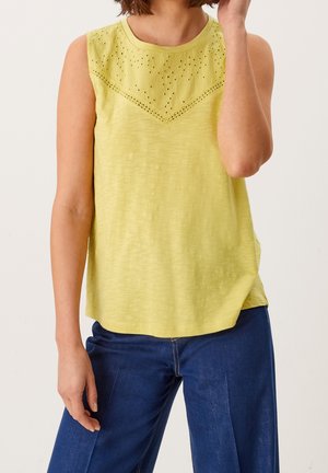 Żółta bluzka bez rękawów z teksturowanym wykończeniem dziurek przy dekolcie. Miękki materiał o luźnym kroju, zestawiona z niebieskimi spodniami dżinsowymi.