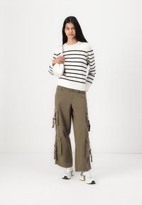 Pull rayé noir et blanc, pantalon cargo olive avec poches et cordons, sac blanc et baskets chunky avec des accents verts.