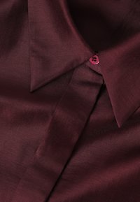 Colletto di camicia in satin bordeaux con patta nascosta e piccolo bottone abbinato visibile vicino al colletto.