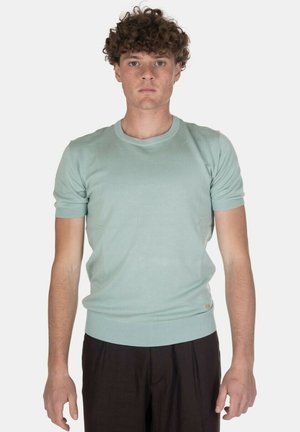Giovane uomo con capelli ricci che indossa una maglietta a maniche corte azzurro chiaro e pantaloni scuri, in piedi davanti a uno sfondo semplice.