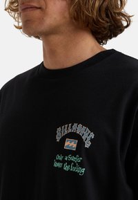 Black cotton t-shirt featuring a graphic print in cream and green, with text "BILLABONG" and "Only a Surfer knows the feeling" below.