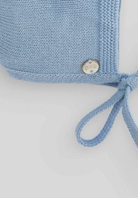 Bonnet en tricot bleu clair avec liens, arborant une étiquette logo argentée marquée "paz" et un détail de couture texturé.