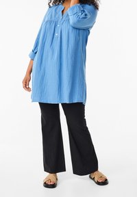 Tunique bleu clair avec un yoke texturé, manches longues et fermeture par boutons. Associée à un pantalon évasé noir et des sandales camel.