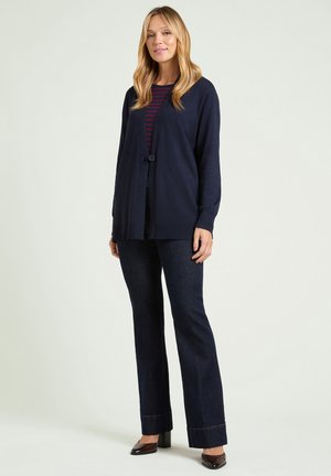 Blond kvinna som bär en marinblå kofta med knappar, röd och marinblå randig topp, mörka jeans och bruna skor med klackar, stående mot en enfärgad bakgrund.