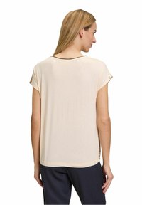 Žena s dlouhými blond vlasy, oblečená v volné, krátkotrvající béžové blůze a černých kalhotách, s zády k bílé pozadí.