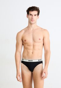 Homme torse nu debout, portant un slip noir avec une ceinture blanche portant la marque "BOSS", devant un fond clair uni.