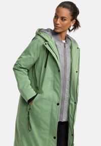 Light green long coat with a hood, featuring black hardware, a zip and snap closure, and layered gray inner jacket with quilting detail.