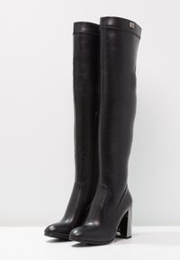 Botas de cuero negro por encima de la rodilla con un alto tacón cuadrado, textura suave y cremallera lateral. Presenta una punta redondeada y costuras decorativas.