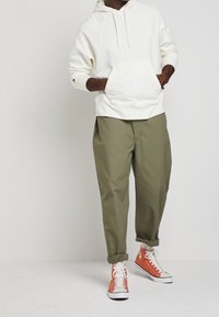 Bílá mikina s kapucí a přední kapsou, v kombinaci s volnými olivově zelenými kalhotami a oranžovými vysokými teniskami. Jednoduchý design a jemné materiály.