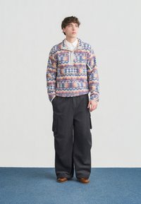 Νεαρός άνδρας στέκεται πάνω σε μπλε χαλί φορώντας πολύχρωμο φλις πουλόβερ με σχέδια, λευκό πουκάμισο με γιακά, φαρδιά μαύρη παντελόνια τύπου cargo και καφέ παπούτσια.