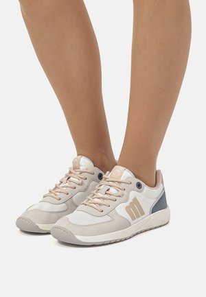 Par de zapatillas beige y blancas con cordones rosa claro usadas sobre piernas bronceadas con un fondo blanco liso.