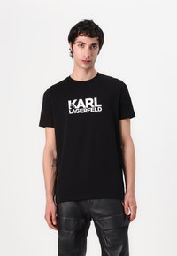 T-shirt en coton noir avec un logo blanc "KARL LAGERFELD" sur la poitrine. Design à col rond. Porté avec un pantalon en cuir noir.