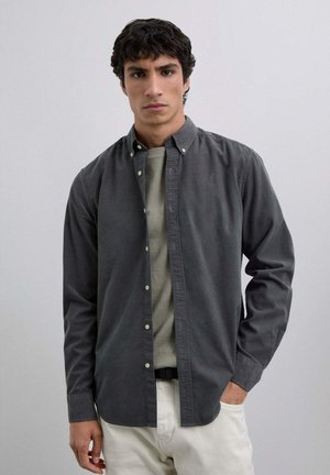 Camisa de botón en color carbón con un acabado suave y texturizado, combinada con una camiseta gris clara por debajo y pantalones color crema.