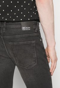 Crne traperice od denima s stražnjim džepom i pravokutnom oznakom s natpisom "DENIM TOM TAILOR." Imaju glatku teksturu i prilagođen dizajn.