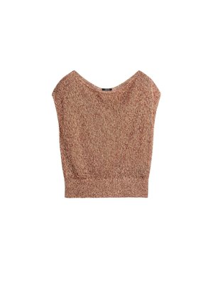 Frau trägt ein braunes, strukturiertes One-Shoulder-Top, einen cremefarbenen asymmetrischen Rock und schwarze Sandalen, steht an einer schlichten weißen Wand.