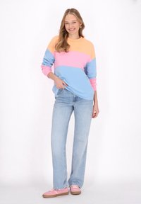 La felpa color block presenta sezioni arancioni, rosa e blu; abbinata a jeans azzurri chiari e sneaker rosa, su uno sfondo bianco.