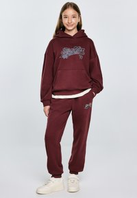 Burgundowa bluza z kapturem z przednią kieszenią i graficznym nadrukiem, w połączeniu z pasującymi dresami z logiem. Całość dopełniają sneakersy.