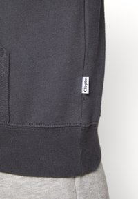Sudadera gris oscuro con dobladillo y puños acanalados, con una pequeña etiqueta blanca "Clepto" cosida en el costado. Textura suave.