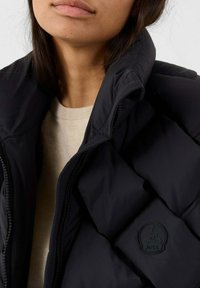 Close-up van een persoon in een zwarte gewatteerde jas over een beige trui, waarbij de hoge kraag van de jas en het logo-patch zichtbaar zijn.