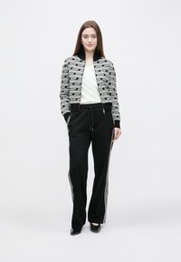 Black and white zippered jacket with repeating logo pattern, paired with black pants featuring side stripes, worn over a white shirt.