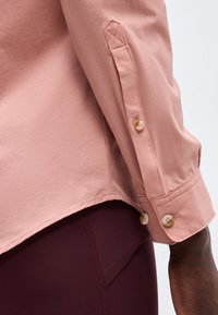 Közeli kép egy személyről, aki világos rózsaszín, gombos mandzsettás hosszú ujjú inget és sötét bordó nadrágot visel.