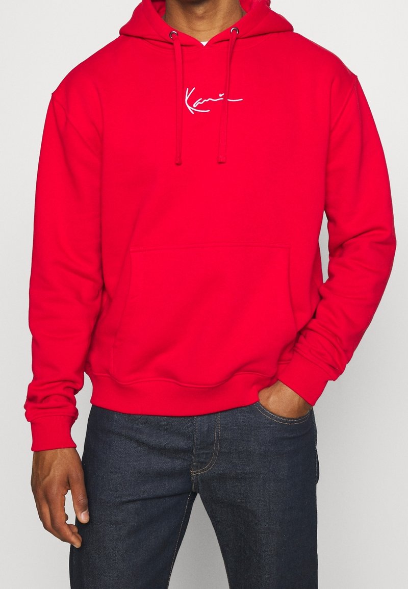 Homme portant un sweat à capuche rouge vif avec poche frontale et logo en écriture blanche, associé à un jean bleu foncé.