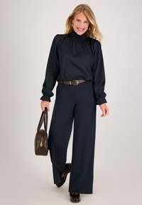 Navy blouse met een gesmokte halslijn en lange mouwen, gecombineerd met wijde navy broek. Een bruine riem en handtas zorgen voor contrast, met glanzende zwarte schoenen.