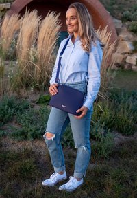 Marineblaue Umhängetasche mit glatter Textur, abgerundeter Form und silberner Hardware, gehalten von einer Person in einem hellblauen Shirt und zerrissenen Jeans.
