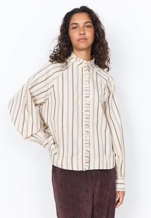 Woman wearing a cream blouse with vertical blue and yellow stripes and ruffle detail down the buttoned front, paired with brown corduroy pants.