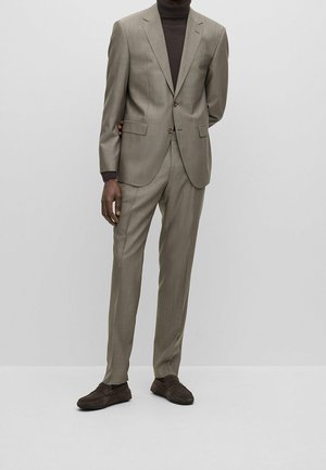 Costume marron clair en tissu texturé, veste à simple boutonnage, deux boutons, poches plaquées, et pantalon assorti. Porté avec un col roulé foncé et des chaussures marron.