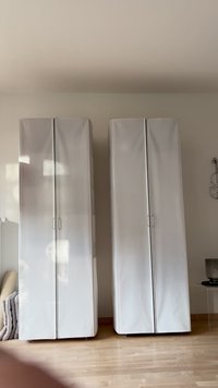 Dvě vysoké, bílé skříně s lesklým povrchem a jednoduchými kovovými úchyty. Každá má dvojité dveře a elegantní, moderní design.