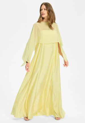 Femme en robe fluide jaune pâle avec manches cape transparentes, regardant sur le côté sur un fond blanc uni.