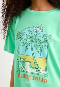 T-shirt vert menthe présentant un design graphique de palmiers, de soleil et de vagues, avec le texte "PARADISE FOUND" en orange sur un fond bleu clair.
