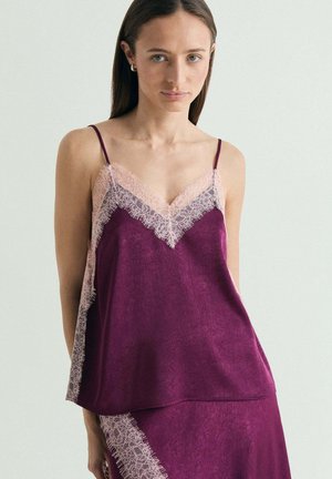 Mujer con camisola de satén morada y falda a juego con ribete de encaje rosa claro, de pie frente a un fondo claro y liso.