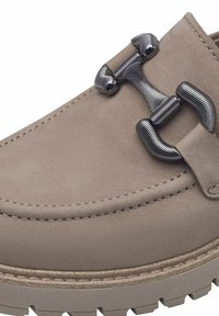 Mocassin en daim taupe avec semelle épaisse et détail en métal argenté en forme de mors sur le dessus, montrant des coutures et textures détaillées.